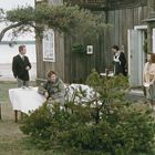 Eine Gruppe von vier Menschen steht in einem Garten vor einem Holzhaus am Seeufer. Ein Tisch ist gedeckt.