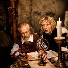 Zwei Männer in historischer Kleidung bei Kerzenlicht, mit Sternenkarte und Armillarsphäre auf einem Tisch ausgebreitet.