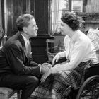 Ein älterer Mann und eine Frau im Rollstuhl halten Händchen und lächeln sich in einem gemütlichen Raum an.