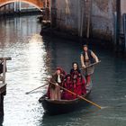 Menschen in historischer Kleidung auf einer Gondel in einem venezianischen Kanal, umgeben von Backsteinbauten und Brücke.