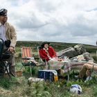 Drei Personen draußen beim Picknick: Einer grillt in Uniform, zwei sitzen entspannt. Tisch mit Decke, Felslandschaft im Hintergrund.