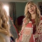 Eine Frau mit blonden Haaren hält eine Holzfigur von Santa Claus, während sie mit einer anderen Person spricht. Weihnachtliche Dekoration im Hintergrund.