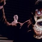 Ein maskierter Charakter mit ungewöhnlicher Maske, düstere Beleuchtung, Hintergrund mit Jalousien und einem gläsernen Kopf.