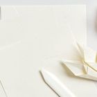 Weiße Papierbögen, ein Falzbein und zwei gefaltete Origami-Herzen auf einer weißen Fläche.