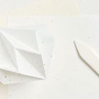 Ein weißes, geometrisches Ornament aus Papier liegt auf mehreren Blättern, daneben ein weißer, länglicher Gegenstand.