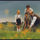 Ein Junge und ein Mädchen mit Brille auf einer Wiese, begleitet von zwei Männern. Der Himmel ist bewölkt.