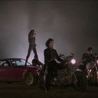 Keine Texte vorhanden. Mehrere Motorräder und ein rotes Auto im Vordergrund. Menschen umgeben die Fahrzeuge, einer steht auf dem Auto.