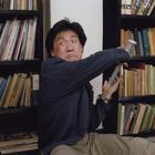 Ein Mann sitzt mit einem erschrockenen Gesichtsausdruck und einem Buch in der Hand vor Bücherregalen.