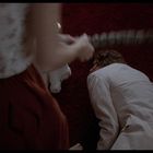 Kein Text erkennbar. Eine Person liegt auf einem roten Teppich, während eine andere Person mit einem Gegenstand daneben steht.