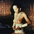 Es gibt keinen sichtbaren Text. Eine Person bindet einen traditionellen Rock, im Hintergrund sind gemusterte Stoffe sichtbar.