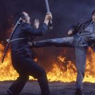 Ein Kampf vor einer großen Feuerwand: Eine Person tritt eine andere, die mit einem Schwert verteidigt.