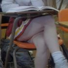 Ein Mensch sitzt auf einem orangefarbenen Stuhl, liest ein Buch, trägt einen rot-weiß-gelben Rock und Turnschuhe.