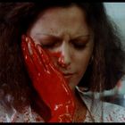 Eine Frau mit geschlossenen Augen hält sich die linke Wange. Ihre Hand und ihr Gesicht sind mit roter Farbe bedeckt.