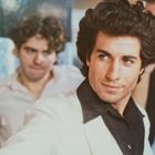 Ich kann keine spezifischen Personen erkennen. Ein Mann trägt ein weißes Jackett und lächelt. Zwei Männer im Hintergrund.