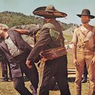 Ein Mann mit großem Hut schlägt einen älteren Mann. Ein anderer Beobachter steht daneben in zerrissener Kleidung.