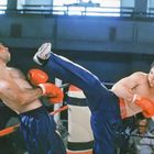 Ein Boxkampf im Ring: Zwei Kämpfer mit roten Handschuhen, einer tritt den anderen, beide tragen blaue Hosen.