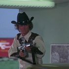 Eine Person in Cowboykleidung mit schwarzem Hut hält eine Waffe und steht in einem Raum. Links ist ein roter Güterzug zu sehen.