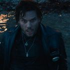 Ein Mann mit nassem Haar und Lederjacke kniet erschrocken im flachen Wasser, seine Halsketten sind sichtbar.