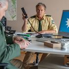 Text: "Polizei" auf Uniformärmeln. Zwei Polizisten, einer hebt den Telefonhörer. Computer zeigt Polizeilogo. Büro mit Karten.