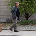 Ein Mann geht mit einem kleinen rauhaarigen Hund an einer Hausfassade und Pflanzen vorbei.