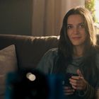 Eine Person sitzt lächelnd auf einem Sofa, hält ein Smartphone. Die Umgebung wirkt gemütlich und warm beleuchtet.