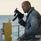 Ein Mann in einer Jeansjacke hält eine Waffe. Er steht draußen, im Hintergrund ist das Meer.
