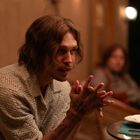 Eine Person sitzt an einem Tisch, die Hände gefaltet, mit einem nachdenklichen Ausdruck. Im Hintergrund ist eine weitere Person unscharf.