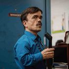 Ein Mann in blauem Hemd hält ein Telefonhörer. An einer Tür steht "MANAGER". Schränke befinden sich im Hintergrund.