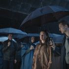Eine Gruppe von Menschen steht im Regen mit Regenschirmen. Ein Mann hält den Schirm für eine Frau hoch. Es ist dunkel und regnerisch.