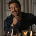 Ein Mann in dunklem Hemd lächelt entspannt, stützt Kinn auf Hand. Im Vordergrund Kerzenlicht und Weingläser.