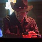 Ein Mann mit Cowboyhut und Lederjacke sitzt an einem Tisch, das Licht ist schwach und neongrell.