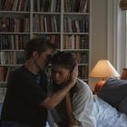 Ein Bücherregal voller Bücher. Zwei Personen sitzen eng umarmt auf einem Bett. Rechts steht eine Tischlampe.