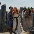 Zwei Personen in altertümlicher Kleidung stehen zwischen hohen Steinstatuen, umgeben von Frauen mit Blumenkränzen im Haar.