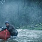 Zwei Personen schieben ein rotes Kanu durch einen flachen, strömenden Fluss in einem nebligen, bewaldeten Gebiet.