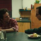 Zwei Männer sitzen in einem Diner und unterhalten sich. Auf dem Tisch sind Salz- und Pfefferstreuer und ein Dessert.