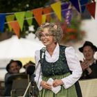 Eine lächelnde Frau in traditioneller Kleidung spricht in ein Mikrofon, im Hintergrund bunte Wimpel und klatschende Personen.