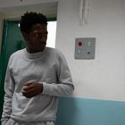 Keine sichtbaren Texte. Ein Mann in grauem Trainingsanzug steht vor einer weißen Wand mit Schalterplatte.