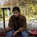 Eine Person sitzt auf einer roten Bank an einer Bushaltestelle, hält einen Stock, umgeben von Bäumen im Hintergrund.