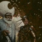 Zwei Personen in extravaganten Kostümen, eine mit weißem Hutturm, die andere in goldener Rüstung, stehen in blumiger Umgebung.