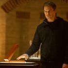 Keine Texte sichtbar. Ein Mann in dunkler Kleidung steht in einem beleuchteten Raum und berührt ein Objekt in einer Holzkiste.