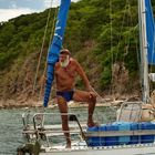 Text: "Der PARADIESJÄGER". Ein älterer Mann in blauer Badehose steht auf einem Segelboot vor grüner Küste.