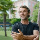 Keine Texte sichtbar. Ein Mann in einem grünen Park, umgeben von Palmen und einem großen Gebäude im Hintergrund.