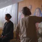 Eine Frau sitzt, während ein Mann ein großformatiges Gemälde mit einer blauen verhüllten Figur bemalt.