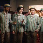 Vier Männer in gestreiften Anzügen und Strohhüten singen in einem festlich geschmückten Raum.