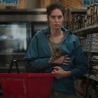 In einem Supermarkt, ein Schild zeigt "BANANAS". Eine Frau in einer blauen Jacke hält ein Kätzchen und einen roten Korb.