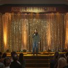 „Festival der Talent*innen“ steht über einer goldenen Bühne, auf der eine Person singt. Publikum schaut zu.