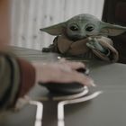 Kleines grünes Wesen mit großen Ohren lehnt auf einem Tisch. Eine unscharfe Hand greift nach einem Knopf.