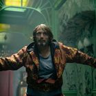 Ein Mann mit Bart stützt sich an grünen Wänden ab, trägt eine reich gemusterte Jacke, im Hintergrund bunte Fenster.