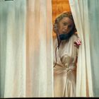 Eine Frau schaut durch einen Spalt in weißen Vorhängen, trägt einen beigefarbenen Morgenmantel.