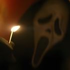 Ein schwarzer Handschuh hält ein brennendes Streichholz vor einer geisterhaften Maske mit offenem Mund und großen Augen.